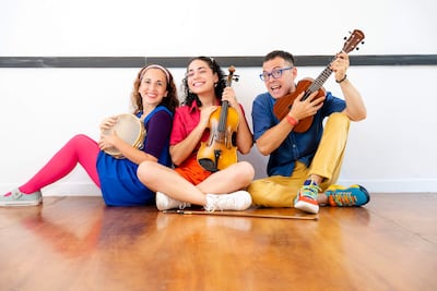 Mujeres con vestido azul y rizado sosteniendo un violín y un pandero, mientras hombre con camisa azul toca ukelele, todos sonrientes.