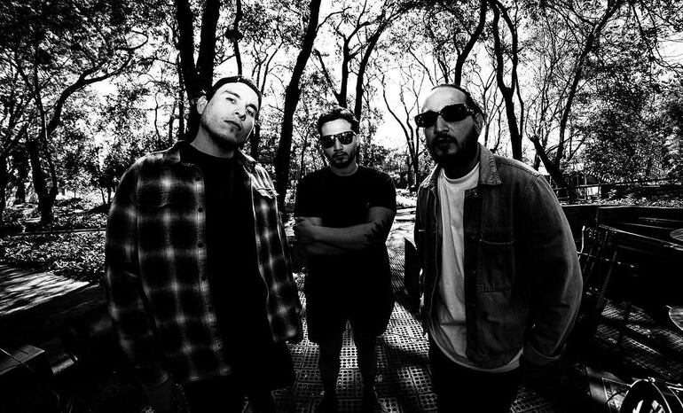 Tres hombres, uno con camisa de cuadros, otro en camiseta negra y el tercero con chaqueta clara, posan en un entorno natural.