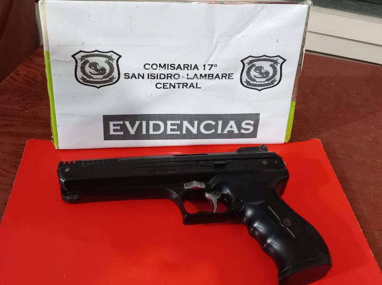 Arma de fuego negra sobre superficie roja, con caja blanca de la Policía Nacional detrás que muestra el escudo y la palabra 'EVIDENCIAS'.