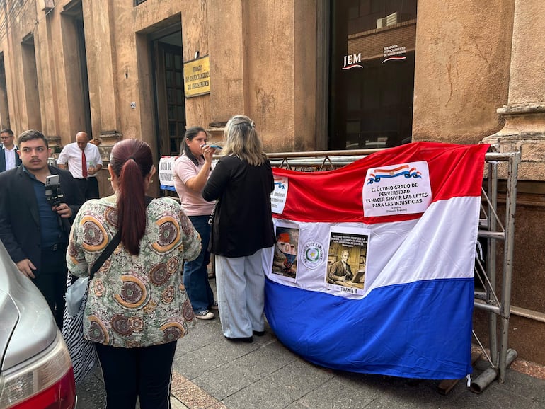 Mujeres conversan y sostienen un vaso, en medio de un ambiente tenso con un letrero crítico sobre el sistema judicial.
