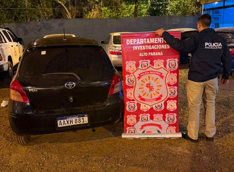 Hombre de espaldas con chaqueta de Policía Investigaciones, sosteniendo cartel en un entorno policial con vehículos en el fondo.