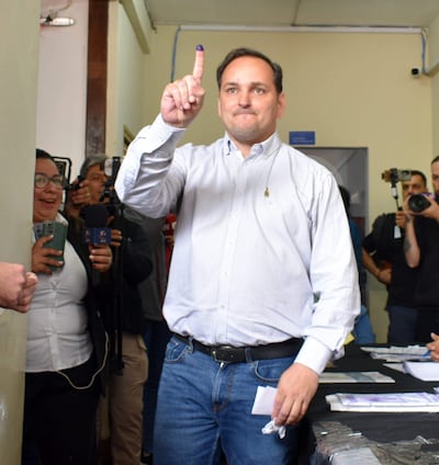 Hombre con camisa de cuadros y jeans levanta dedo índice con tinta en centro de votación, mientras otros observan con sonrisas.