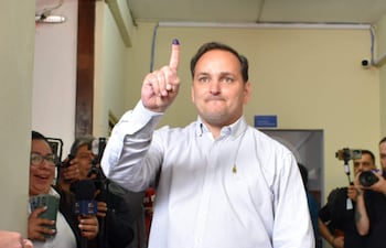 Hombre con camisa de cuadros y jeans levanta dedo índice con tinta en centro de votación, mientras otros observan con sonrisas.