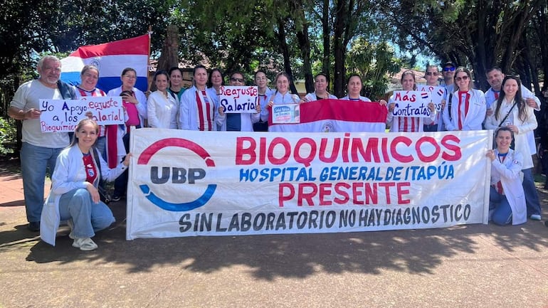 Grupo de bioquímicos en batas blancas sosteniendo pancartas sobre mejora salarial en un entorno soleado.