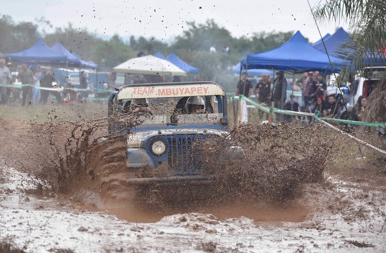Vehículo todoterreno azul del Team Mbuyapey salpicando barro, rodeado de espectadores entusiastas en un ambiente de competición.