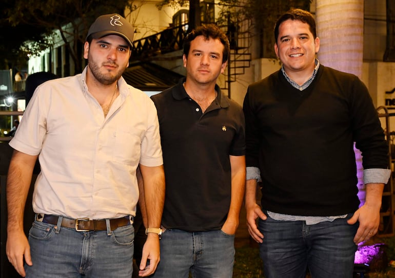 Tres hombres de pie en una calle, uno con camisa blanca y sombrero gris, otro en camiseta negra y el tercero con suéter negro y sonrisa.