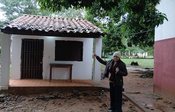 Mujer con gorro de lana y chaqueta negra frente a construcción blanca, señalando una pared. Ambiente natural detrás.