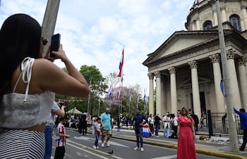 Mujer con top blanco tomando fotos, rodeada de personas, banderas de Paraguay ondeando, y un edificio con inscripción en el fondo.