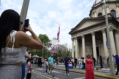 Mujer con top blanco tomando fotos, rodeada de personas, banderas de Paraguay ondeando, y un edificio con inscripción en el fondo.