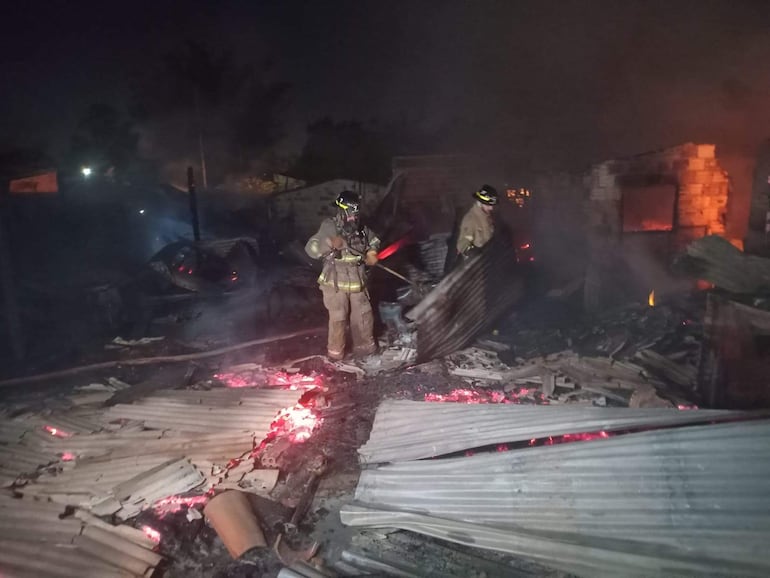 Bomberos en trajes de protección luchan contra un incendio en viviendas, con llamas y humo en la escena.