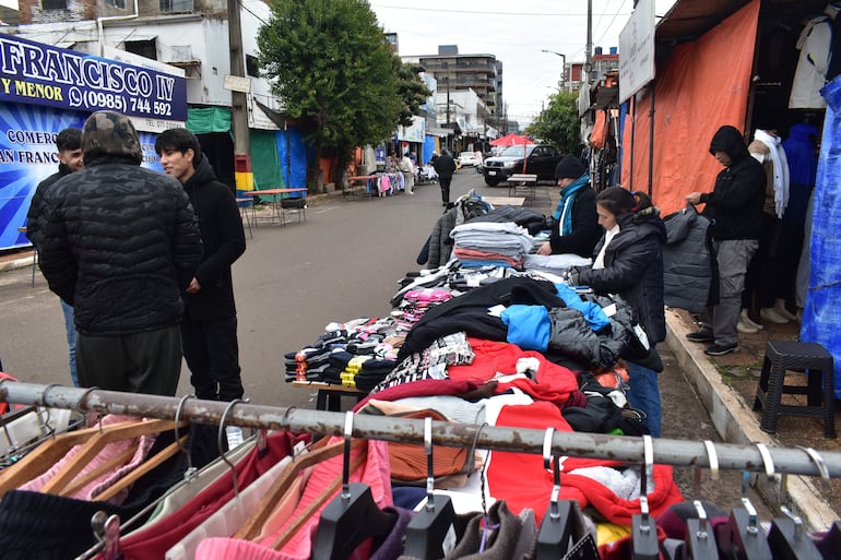 Tres hombres conversan; uno con chaqueta negra. Dos mujeres organizan ropa en mesas, rodeados de un ambiente urbano.
