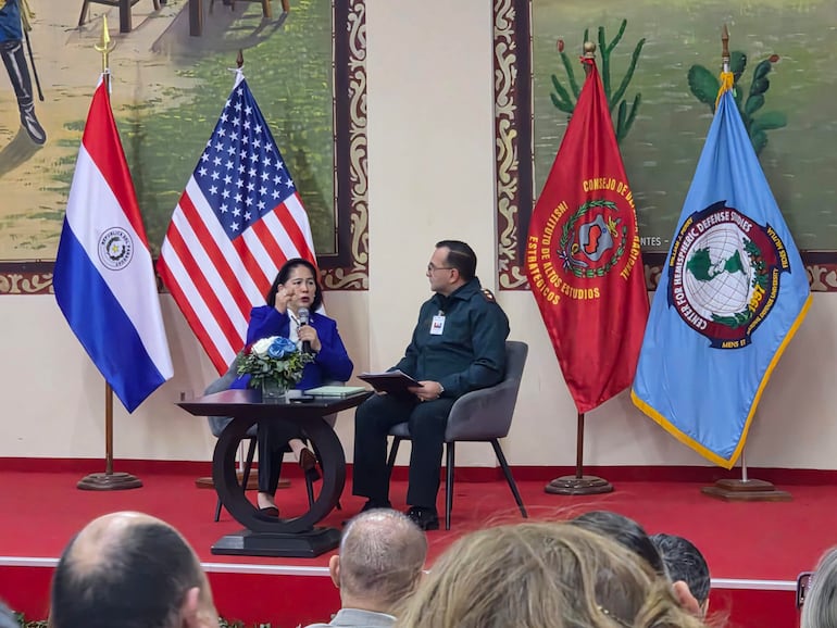 Dra. Celina Realuyo con micrófono en mano, en un escenario decorado con banderas de Paraguay y Estados Unidos, rodeada de asistentes.