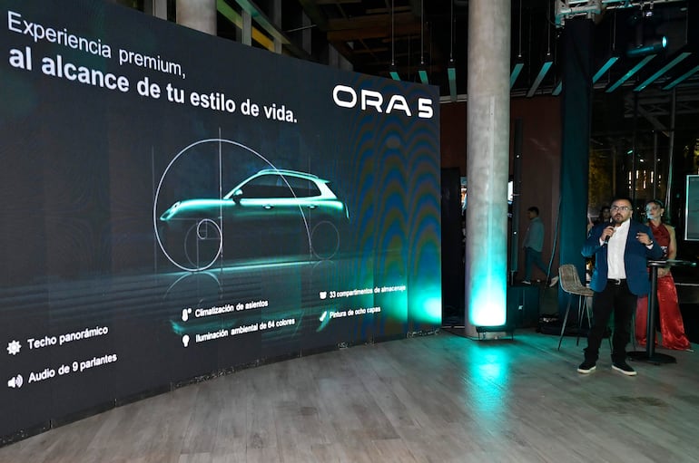 Hombre con traje azul y mujer en vestido rojo en evento formal, presentando el automóvil ORA 5 con pantalla de gráficos detrás.