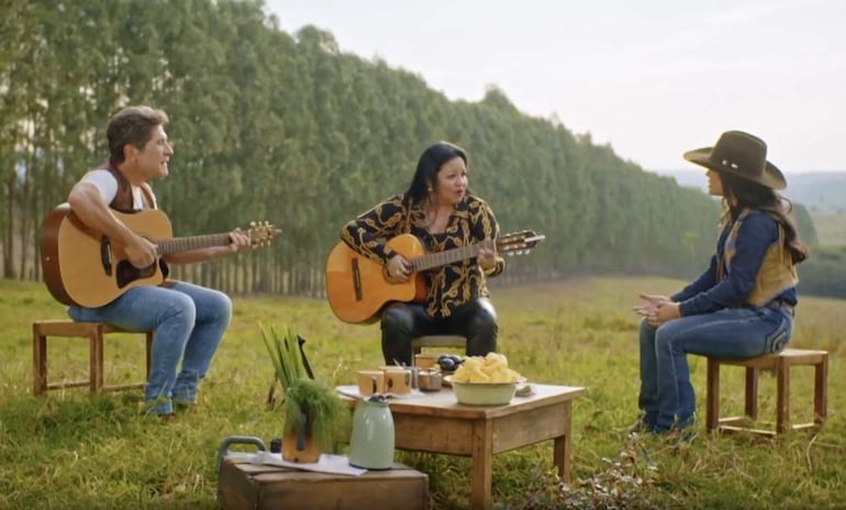 Perla en atuendo decorado y Fiorella en estilo casual con sombrero, sentadas junto a un guitarrista en un entorno natural.