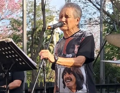 Hombre mayor con cabello canoso canta en micrófono, vestido con camiseta negra con músicos, en un escenario al aire libre con árboles.