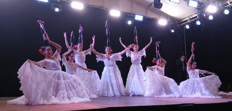 Seis bailarinas en vestidos blancos, perfectamente alineadas, realizando un baile tradicional con entusiasmo en un escenario iluminado.