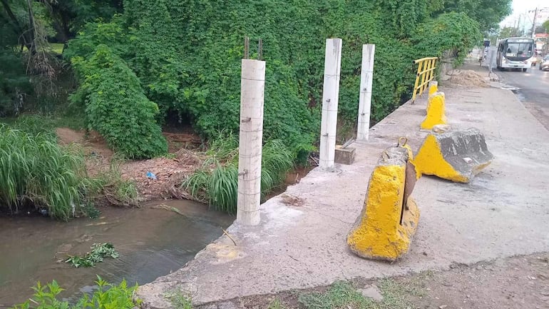Puente en construcción sobre arroyo Mburicaó, sin barandales, con vegetación densa y un vehículo al fondo.