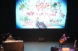 Dos hombres en un escenario oscuro: uno dibuja en una mesa y el otro toca la guitarra, con proyecciones coloridas detrás.
