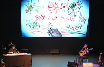Dos hombres en un escenario oscuro: uno dibuja en una mesa y el otro toca la guitarra, con proyecciones coloridas detrás.