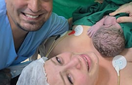 Mujer feliz sonríe con su bebé en brazos, mientras un hombre en camisa azul la acompaña en un ambiente hospitalario.
