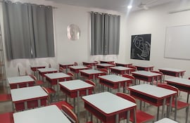 Aula vacía con escritorios blancos y sillas rojas, cortinas grises, y un mapa de países vecinos en la pared.