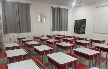 Aula vacía con escritorios blancos y sillas rojas, cortinas grises, y un mapa de países vecinos en la pared.