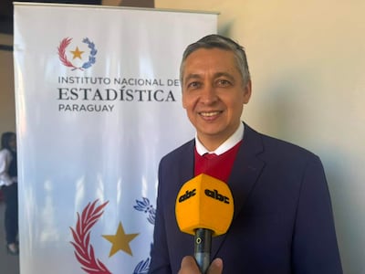 Iván Ojeda sonríe en traje oscuro y suéter rojo, sosteniendo un micrófono en un evento con público atento.