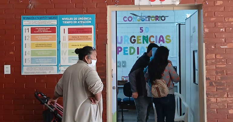 Una mujer con carrito de bebé observa desde el lado izquierdo, mientras otro hombre y mujer esperan en la entrada de Urgencias.
