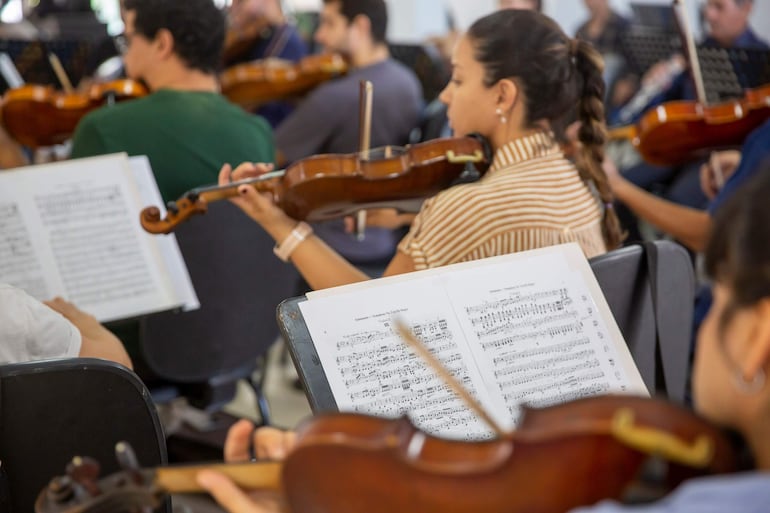Músicos de la orquesta, en su mayoría con violines, muestran concentración mientras ensayan de pie y sentados en un espacio iluminado.