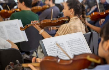 Músicos de la orquesta, en su mayoría con violines, muestran concentración mientras ensayan de pie y sentados en un espacio iluminado.