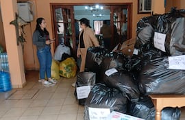 Dos personas en chaquetas, organizando donativos en un espacio con bolsas y cajas, rodeadas de un ambiente sencillo y colorido.