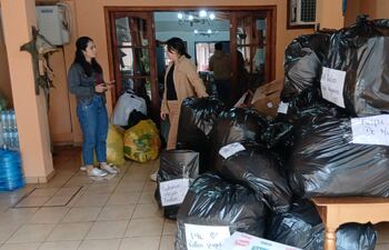 Dos personas en chaquetas, organizando donativos en un espacio con bolsas y cajas, rodeadas de un ambiente sencillo y colorido.