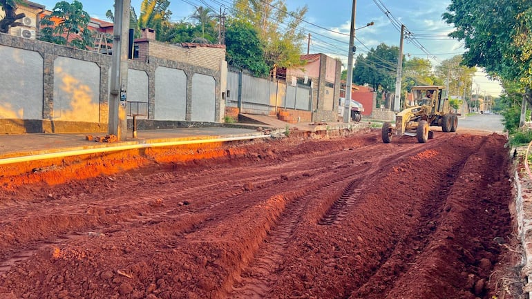Escena de obra en la avenida Avelino Martínez, con máquina motoniveladora trabajando sobre terreno rojo sin personas visibles.