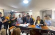Hombre con traje oscuro y corbata sostiene documentos, rodeado de periodistas con cámaras y micrófonos en un ambiente de oficina.