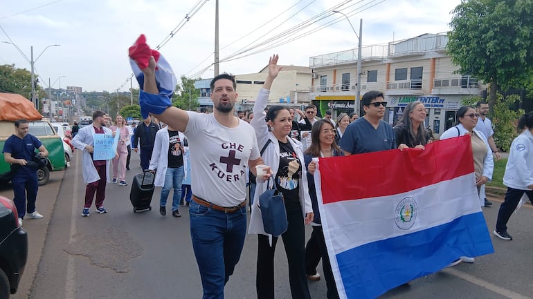 Médicos con batas y scrubs marchan, mostrando carteles y una bandera, en un entorno urbano.