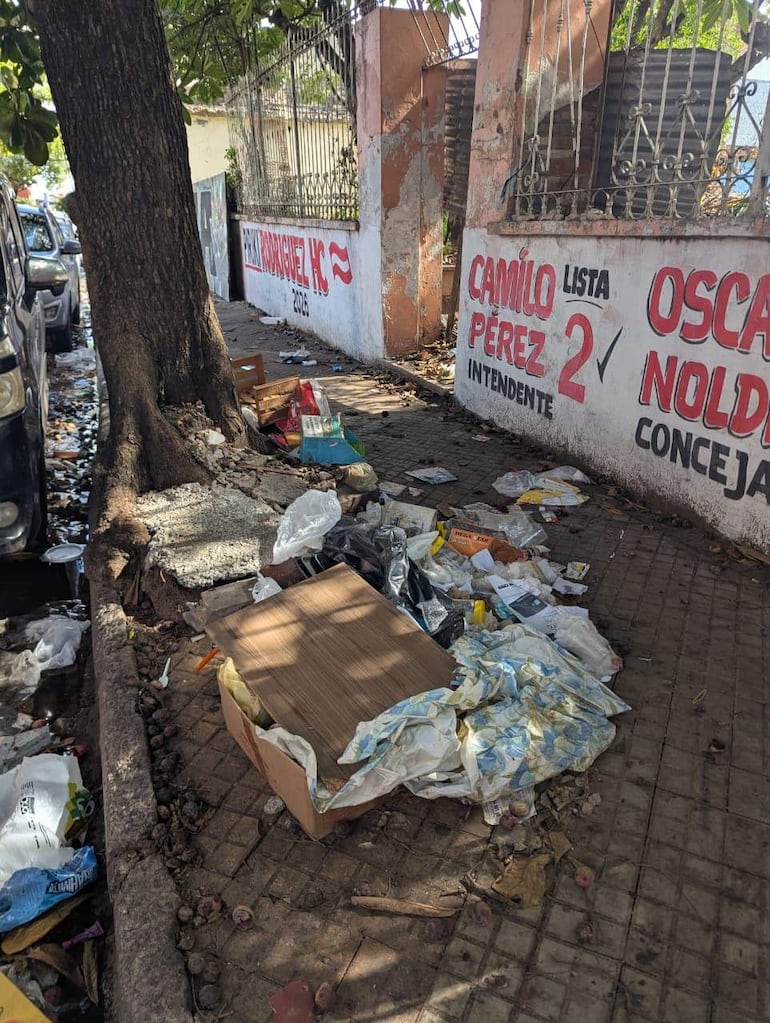 Montón de basura en vereda de ladrillos, sin personas, con grafitis políticos en el fondo.