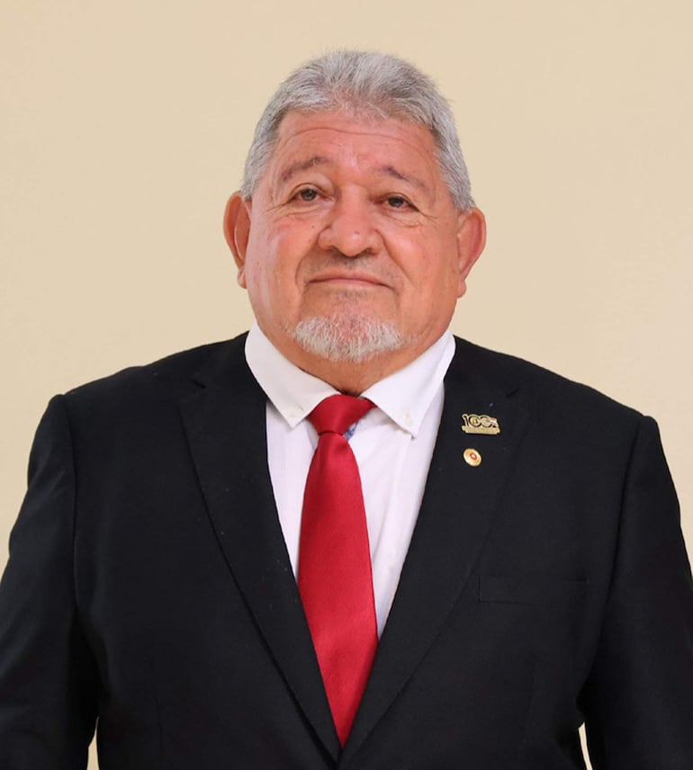 Hombre de mediana edad con cabello canoso, vestido de traje oscuro y corbata roja, posando erguido frente a la cámara en un fondo neutro.