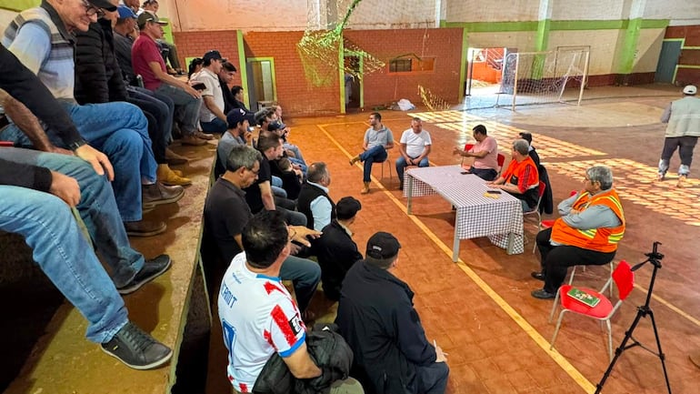 Tres personas en mesa con mantel a cuadros discuten. Un hombre lleva camiseta blanca con detalles rojos, mientras otros escuchan atentamente en un gimnasio.