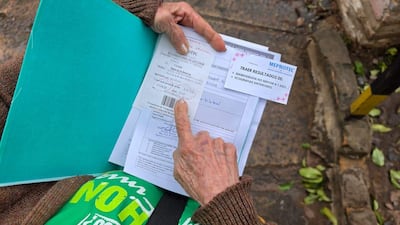 María Gutiérrez de 74 años en suéter marrón y camiseta verde, sostiene documentos médicos en un entorno urbano con pavimento y hojas caídas.