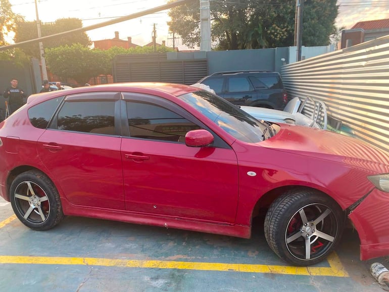 Vehículo rojo en estacionamiento con llanta dañada. El entorno incluye contenedores metálicos y edificios modernos.