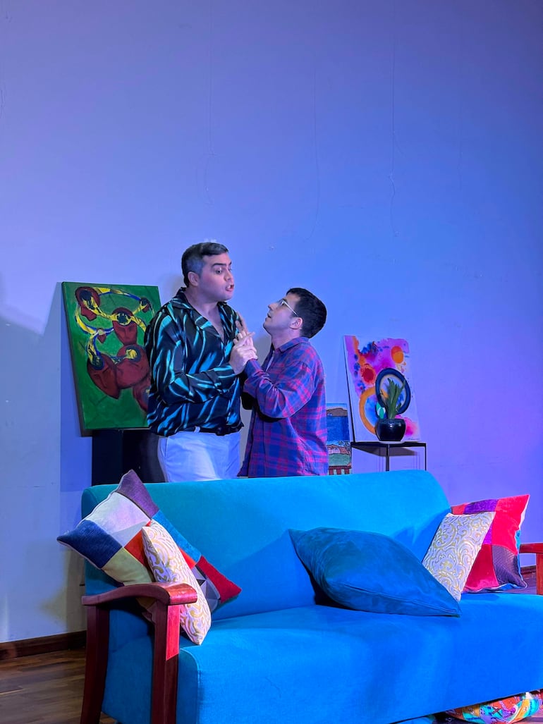 Dos hombres en escena teatral; uno gesticula mientras el otro lo escucha. Ambiente con sofá azul y pinturas decorativas.