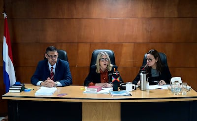 Mujer de cabello rubio con anteojos habla en mesa de juicio, mientras las otras dos personas la escuchan atentas.