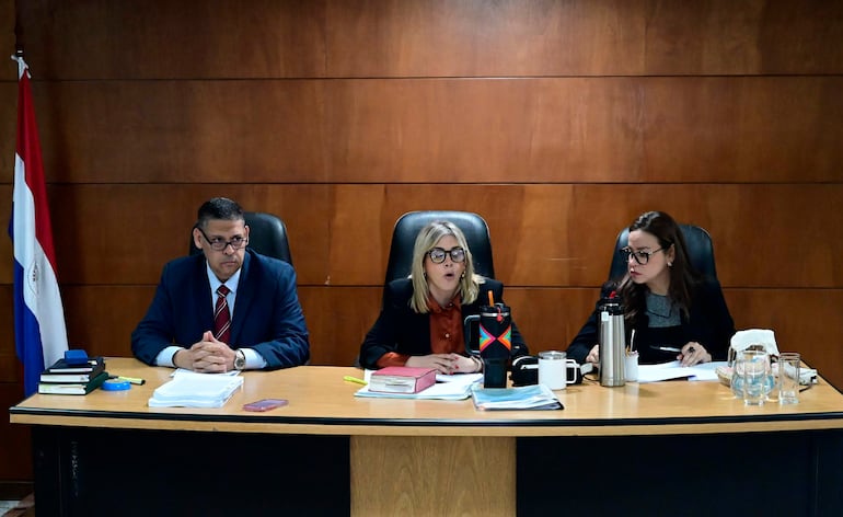 Mujer de cabello rubio con anteojos habla en mesa de juicio, mientras las otras dos personas la escuchan atentas.