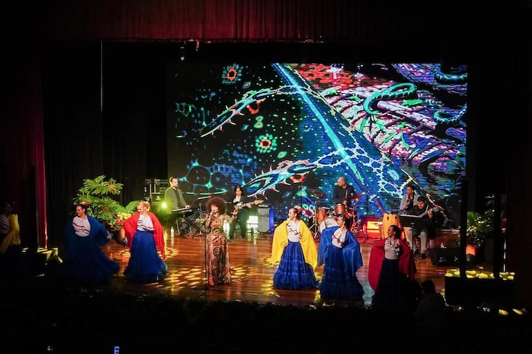 Seis mujeres en trajes tradicionales bailan en el escenario, mientras cinco músicos tocan instrumentos, creando un ambiente festivo y colorido.
