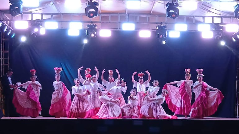 Grupo de bailarinas en trajes tradicionales rosa y blanco, mostrando expresiones felices mientras danzan en un escenario iluminado.