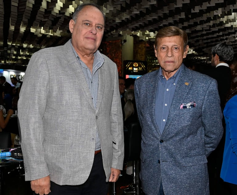Hombre canoso en chaqueta gris y camisa de cuadros junto a compañero en chaqueta azul, ambos con expresiones serias en evento formal.