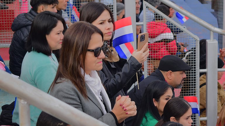 Mujer con gafas de sol sostiene un teléfono, mientras otras personas levantan banderas durante el desfile de independencia.