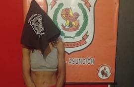 Mujer con rostro cubierto por pañuelo negro en comisaría, vistiendo camiseta sin mangas y pantalones cortos, rodeada de un ambiente policial.