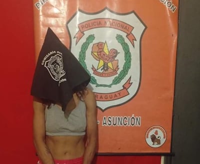 Mujer con rostro cubierto por pañuelo negro en comisaría, vistiendo camiseta sin mangas y pantalones cortos, rodeada de un ambiente policial.