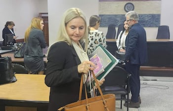 Ana Girala, con carpeta y bolso, se retira de la sala. En el fondo.